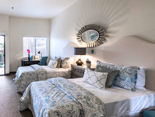 Bright bedroom with two twin beds, patterned bedding, a nightstand with a lamp, and a small desk by a window.