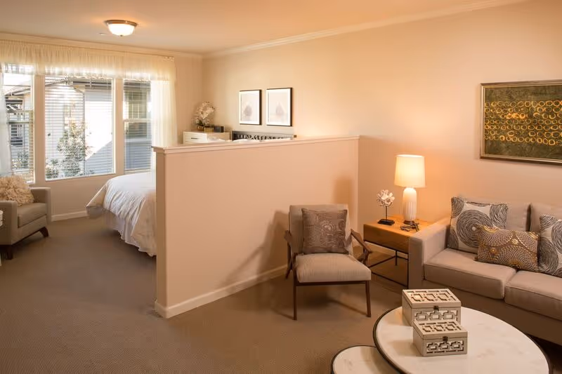 A cozy living space in Oakmont of Fresno featuring a small living room with a beige sofa, decorative pillows, a side table with a lamp, and an armchair. A half wall separates the living area from a bedroom with a bed, dresser, and large window letting in natural light.