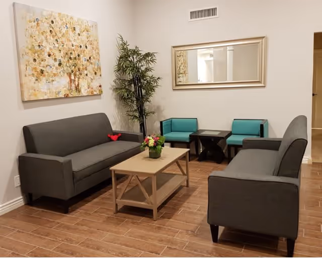 A cozy living room with two gray sofas facing each other, a wooden coffee table with a flower arrangement in the center, two teal chairs with a small black table between them, a tall green plant in the corner, a large abstract painting on the left wall, and a rectangular mirror on the right wall.