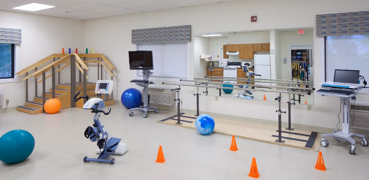 A physical therapy room with exercise equipment including a stationary bike, parallel bars, therapy stairs, exercise balls, and orange cones. There is a large mirror on the wall reflecting a kitchenette area with wooden cabinets and a refrigerator. Two windows with patterned valances let in natural light.