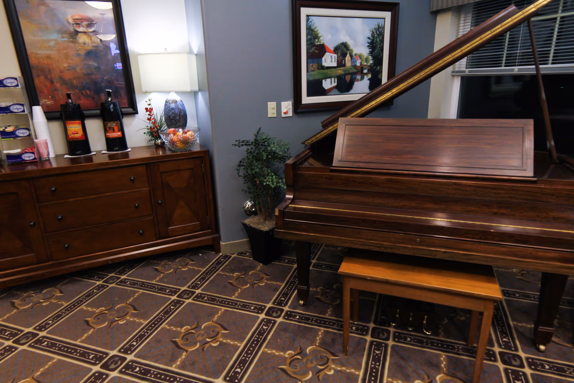A common room featuring a grand piano, a wooden sideboard with coffee dispensers and a lamp, and framed artwork on the walls.