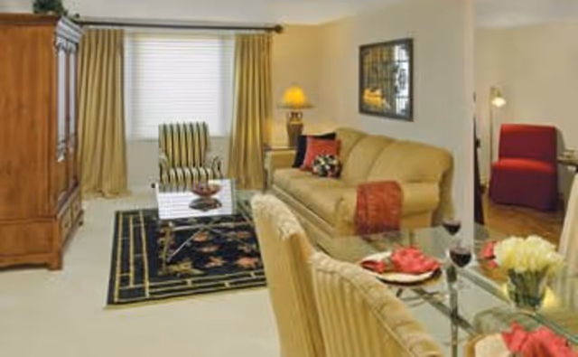 Living room with a beige sofa, striped armchair, glass coffee table on a dark rug, and a nearby glass dining table set for two.