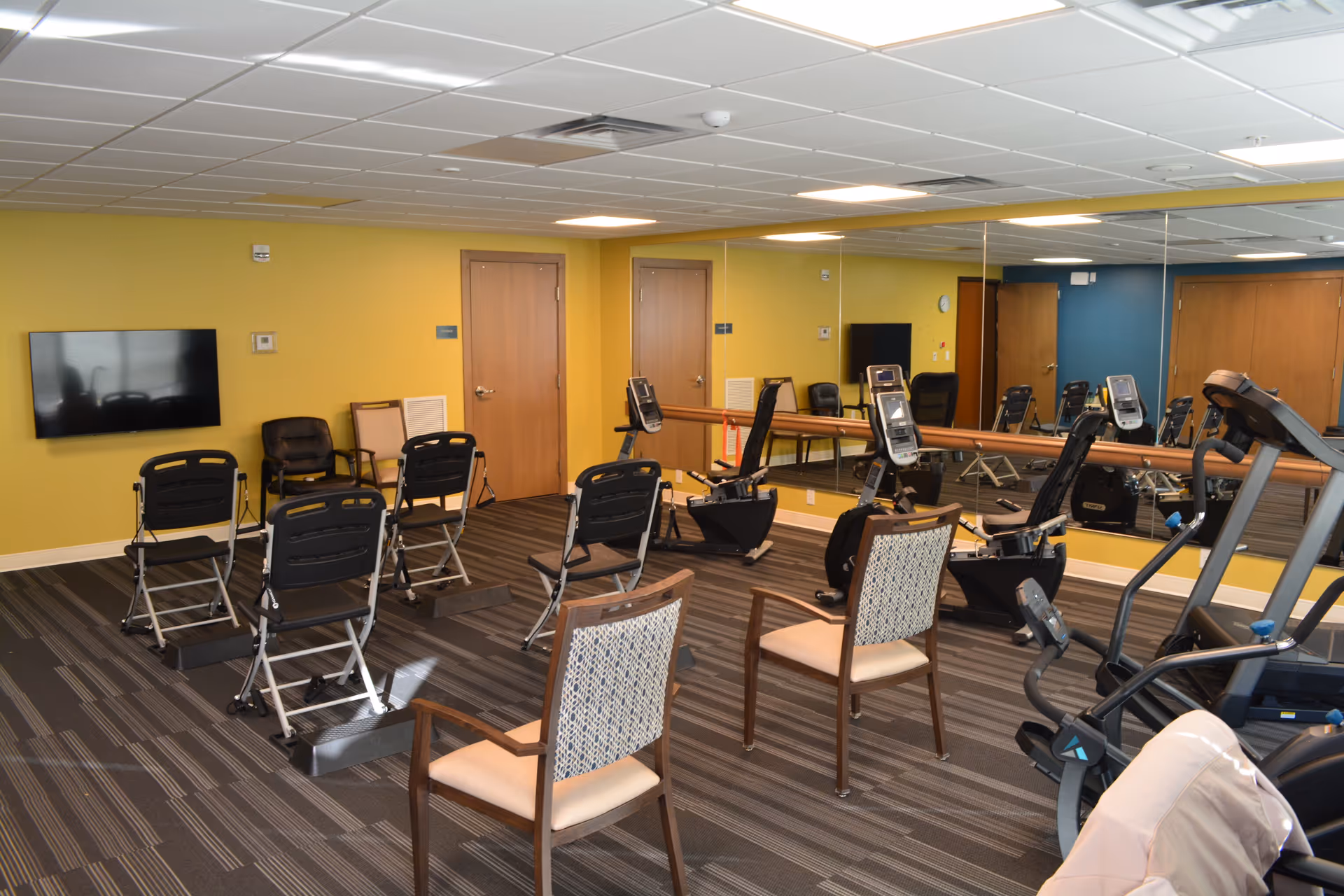 Exercise room with several chairs arranged in rows, two recumbent exercise bikes, a treadmill, a large wall mirror, a wall-mounted TV, and yellow and blue walls with wooden doors.