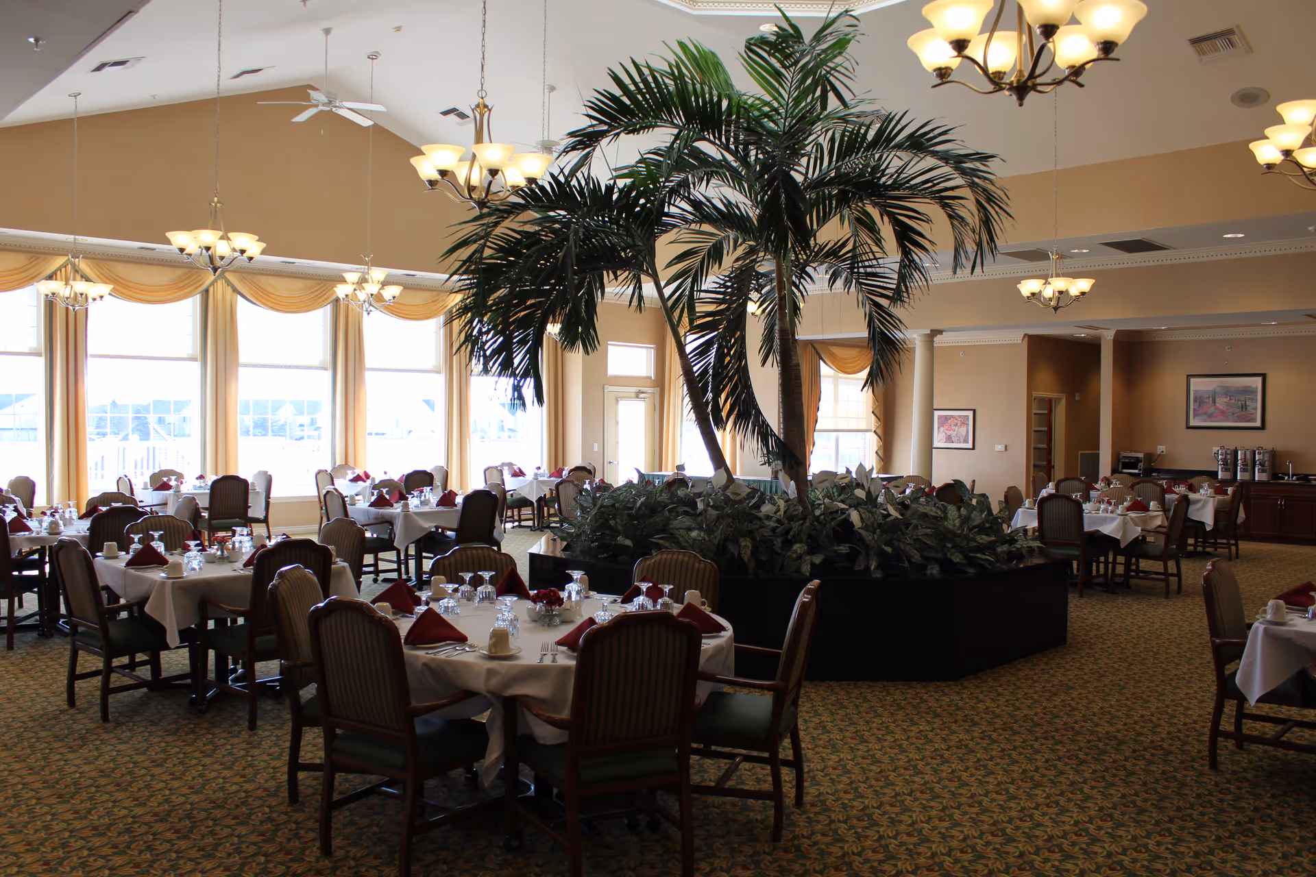 Spacious dining room with multiple round tables covered with white tablecloths, set with glasses, cups, and red napkins. A large indoor palm tree and greenery centerpiece is in the middle of the room. The room has large windows with yellow curtains, chandeliers hanging from the ceiling, and a carpeted floor.