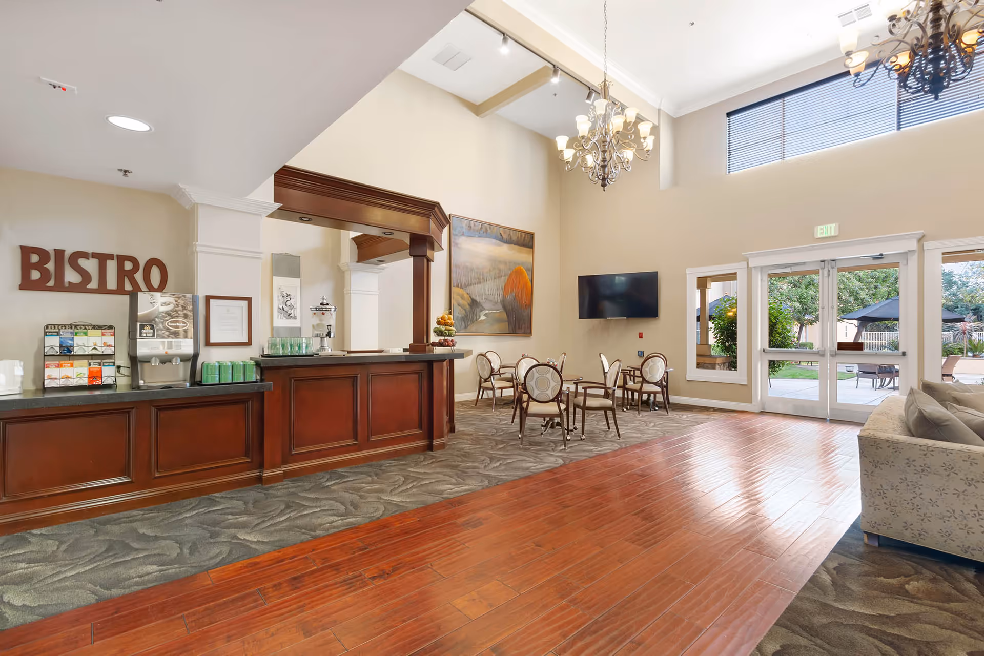 Interior view of a senior living facility's bistro area with a wooden counter, beverage station, small round tables with chairs, a large painting on the wall, a flat-screen TV, chandeliers, and large windows and doors leading to an outdoor patio.