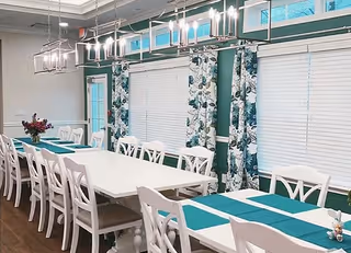Bright dining room with long white tables, teal placemats, chairs, and floral curtains.