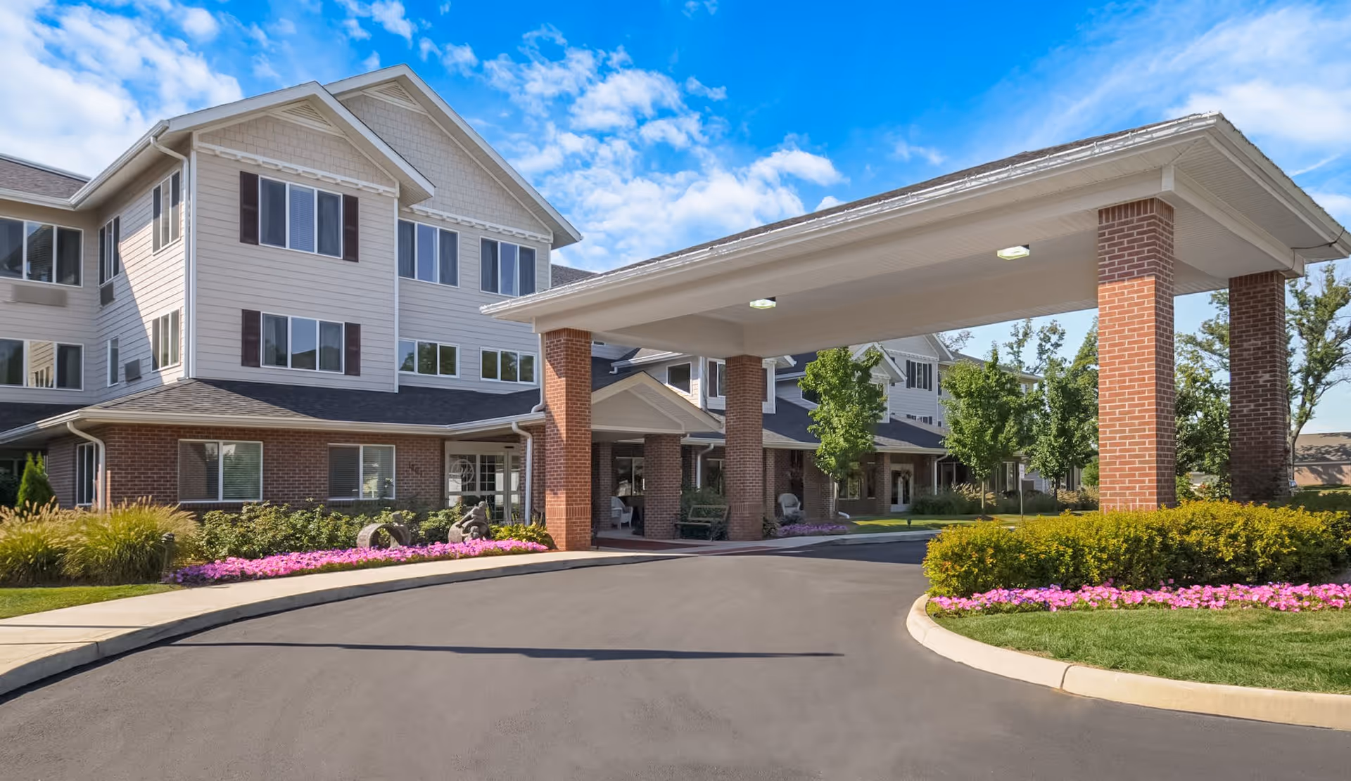 Exterior view of a senior living facility building with a covered entrance supported by brick pillars, surrounded by landscaped greenery and flower beds under a blue sky with some clouds.