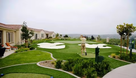 A person playing golf on a putting green at an outdoor area of a senior living facility with single-story buildings and landscaped greenery surrounding the golf area.