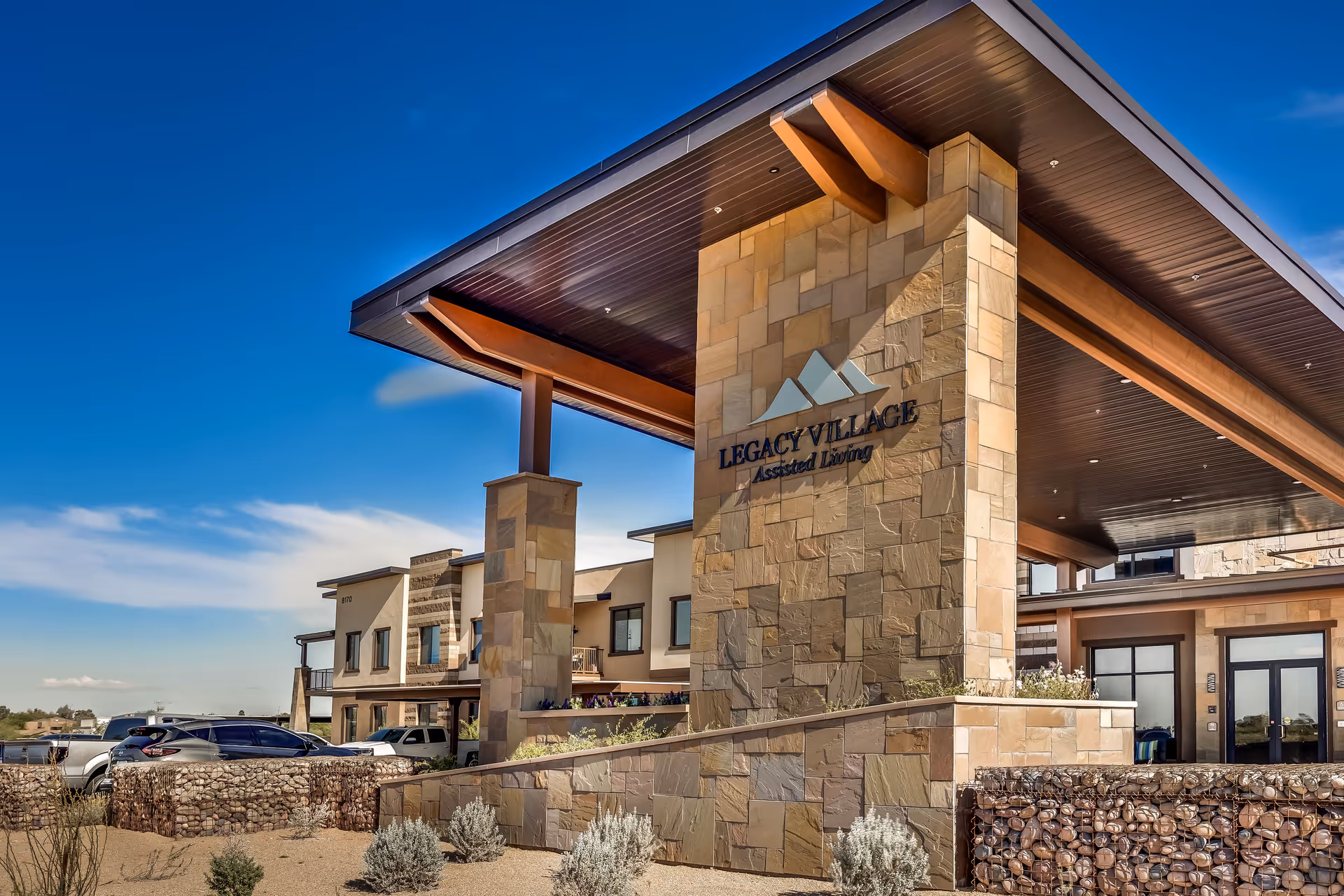Exterior view of Legacy Village Assisted Living facility showing a modern stone and wood entrance with a large covered canopy, clear blue sky, and parked cars nearby.