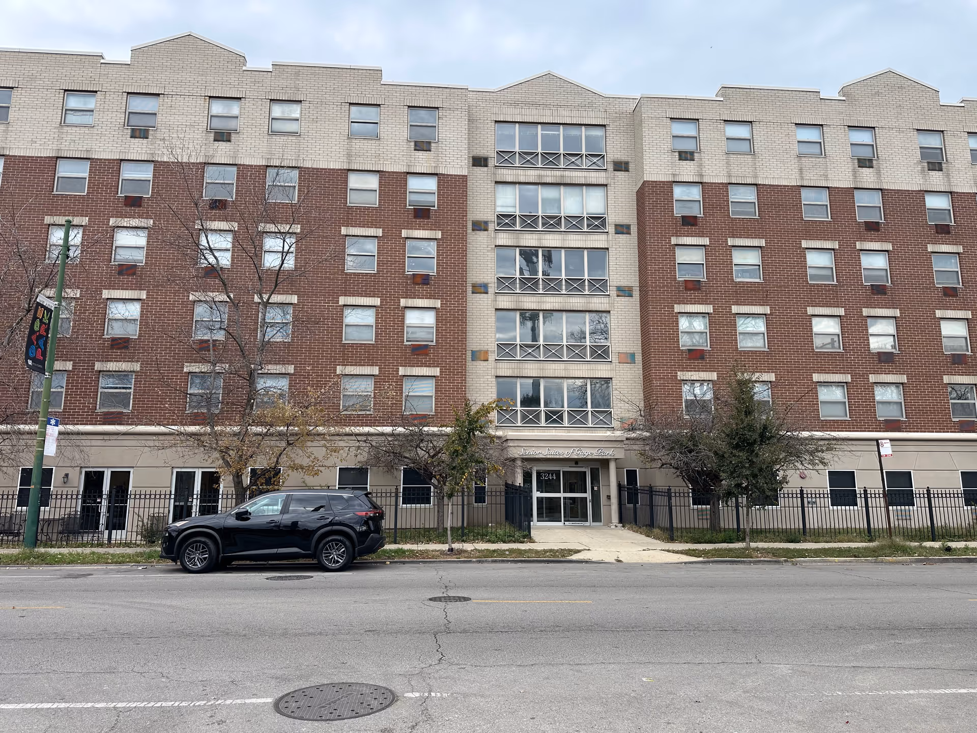 Front exterior view of a multi-story senior living facility named Senior Suites of Gage Park. The building has a combination of beige and red brick facade with multiple windows. A black SUV is parked on the street in front of the building, and there are a few leafless trees and a black metal fence along the sidewalk.