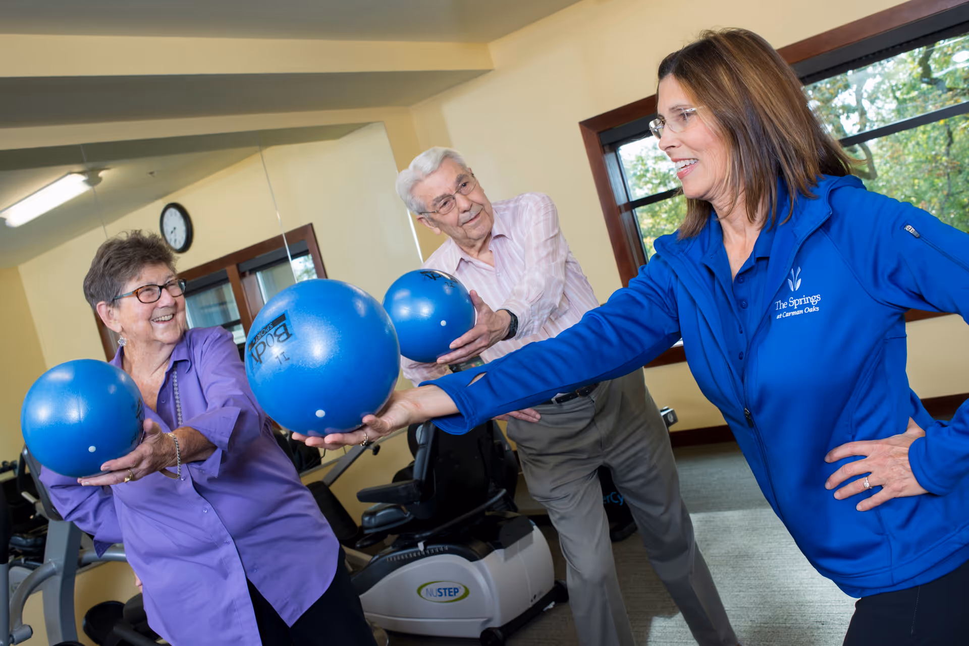 Two elderly individuals, a woman and a man, are participating in a fitness activity with a female instructor in a gym room. They are holding blue exercise balls and stretching their arms forward. The instructor is wearing a blue jacket with The Springs at Carman Oaks logo. Exercise equipment and a large mirror are visible in the background.