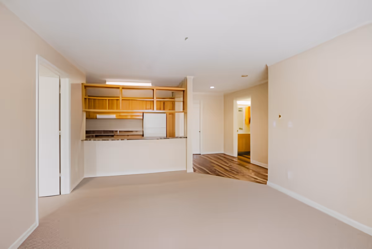 Empty open-plan living room with beige walls and carpet, a pass-through counter revealing a small kitchen with wooden cabinets and a short hallway.