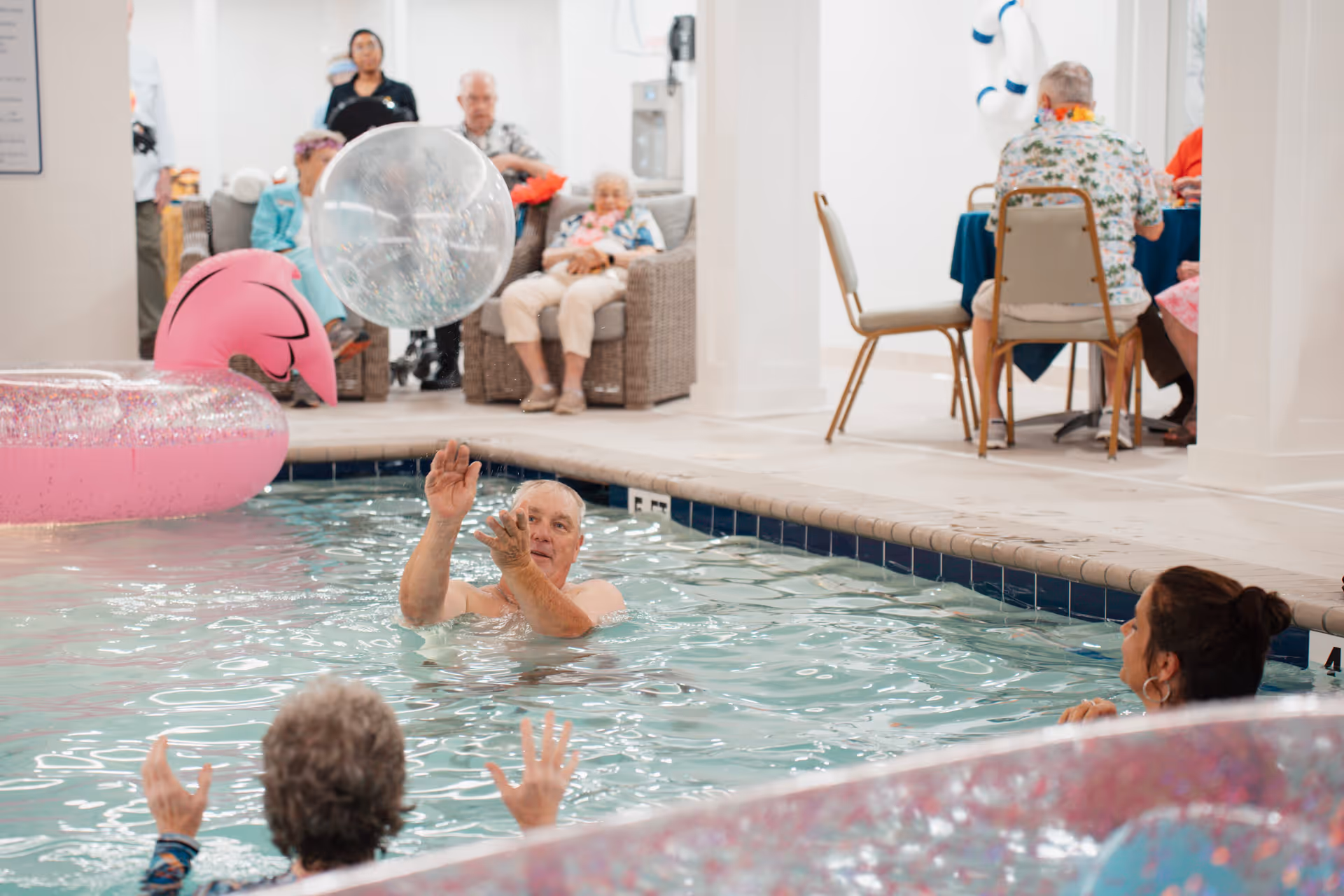 Several elderly people enjoying time in an indoor swimming pool, playing with a large transparent beach ball and a pink inflatable float. In the background, other seniors are seated on wicker chairs and at a table, socializing in a bright, white room.
