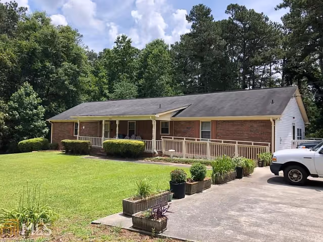 Single-story brick building with a front porch and wheelchair accessible ramp, surrounded by green lawn and trees under a partly cloudy sky. Several potted plants line the driveway, and a white vehicle is parked on the right side.