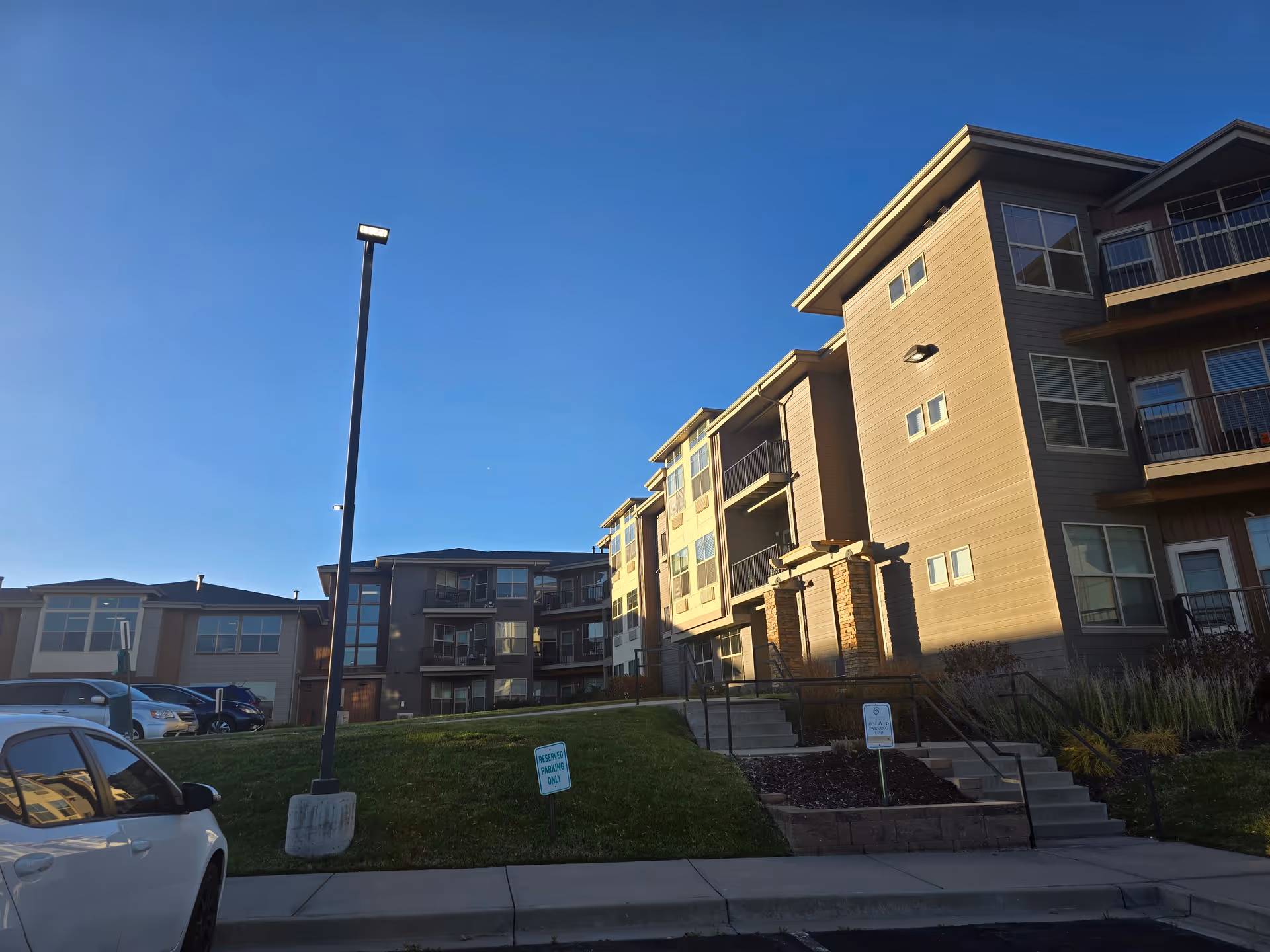 Exterior view of a multi-story residential building with balconies, a grassy area, stairs leading up to the entrance, and parked cars under a clear blue sky.