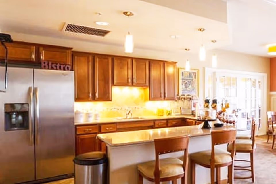 A bright kitchen area with wooden cabinets, a stainless steel refrigerator, a kitchen island with three wooden chairs, pendant lights hanging from the ceiling, and a dining area visible in the background with large windows.