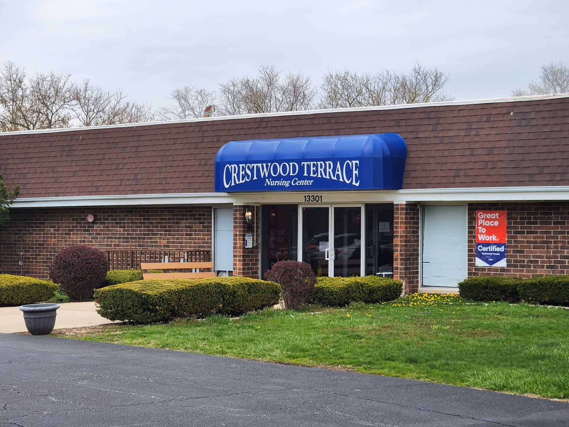 Front entrance of Crestwood Terrace nursing center with a blue awning, brick facade, shrubs, and a small parking area.