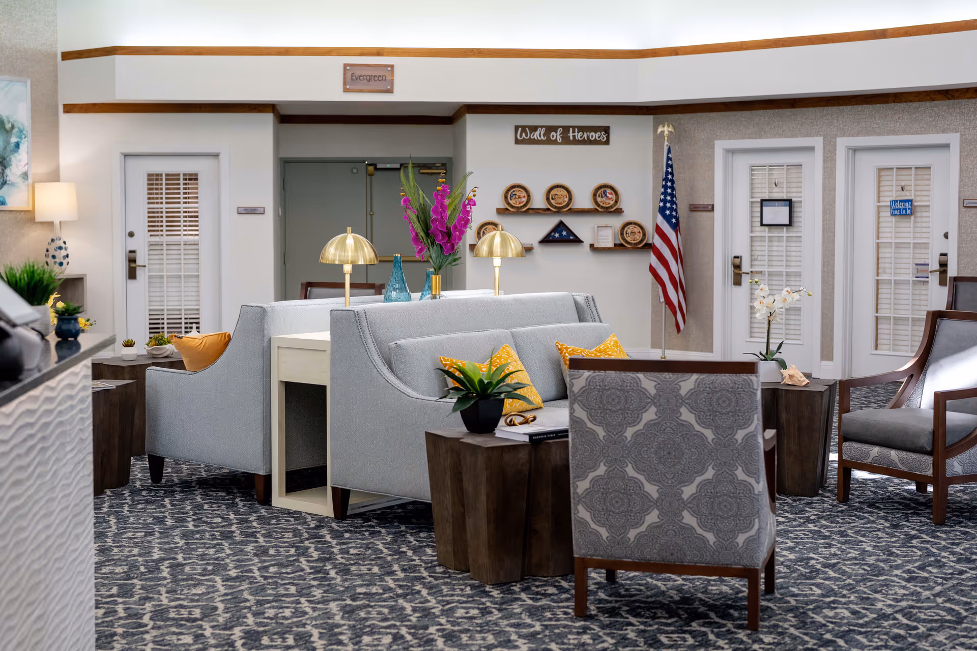 A cozy and well-decorated senior living facility common area with patterned carpet, light gray sofas with yellow pillows, wooden side tables, and brass table lamps. The background features a 'Wall of Heroes' display with plaques and an American flag, along with doors and a lamp on the left side.