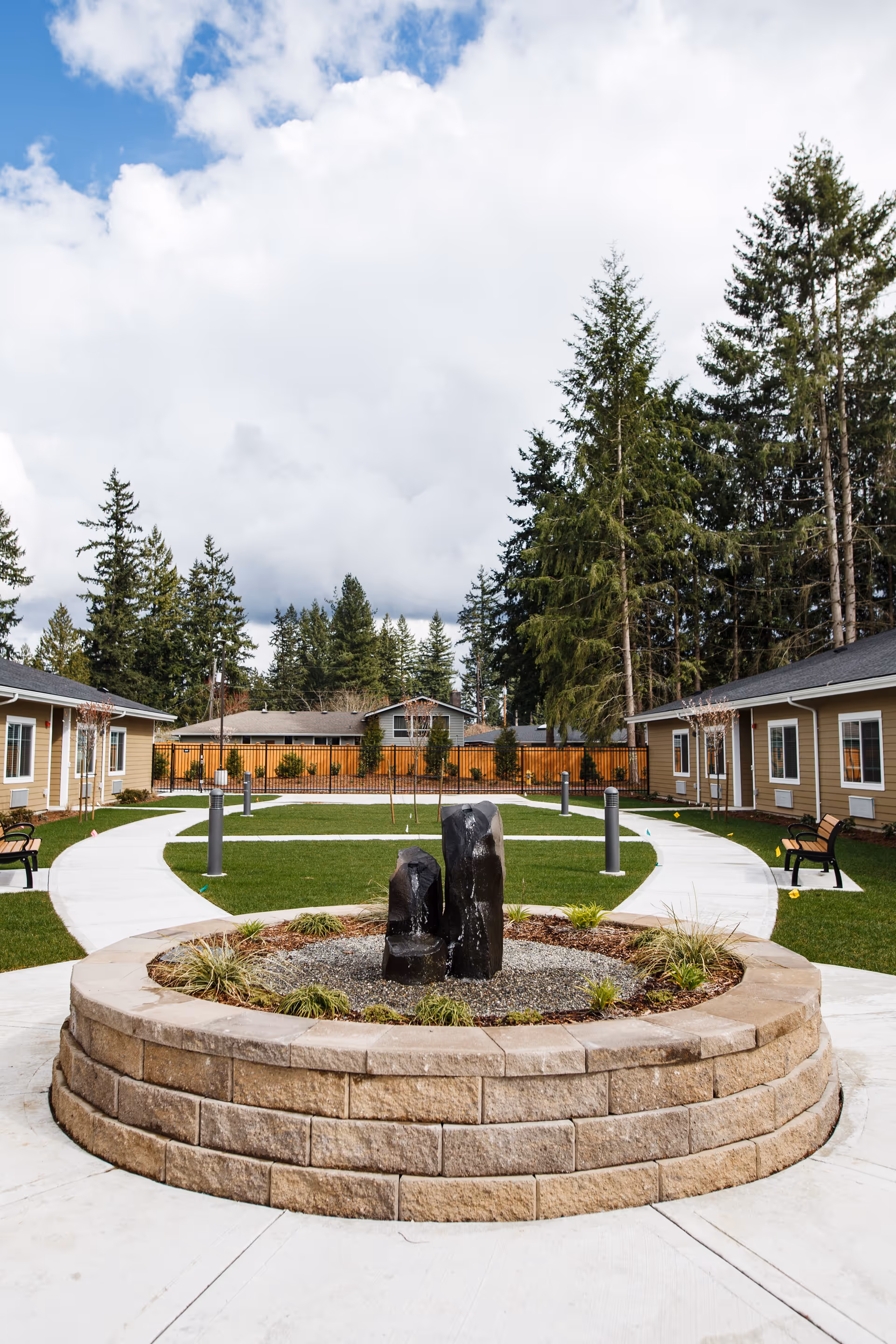 Outdoor courtyard area at Fieldstone Memory Care Marysville featuring a circular stone planter with three black stone water features in the center, surrounded by green grass, paved walkways, benches, and single-story beige buildings with white trim. Tall evergreen trees and a partly cloudy sky are visible in the background.