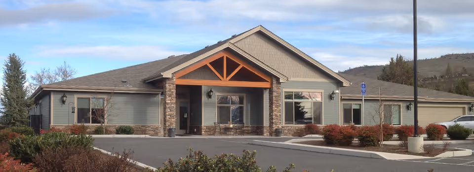 Front exterior of a single-story senior living building with a covered entrance, parking lot, and landscaped shrubs.