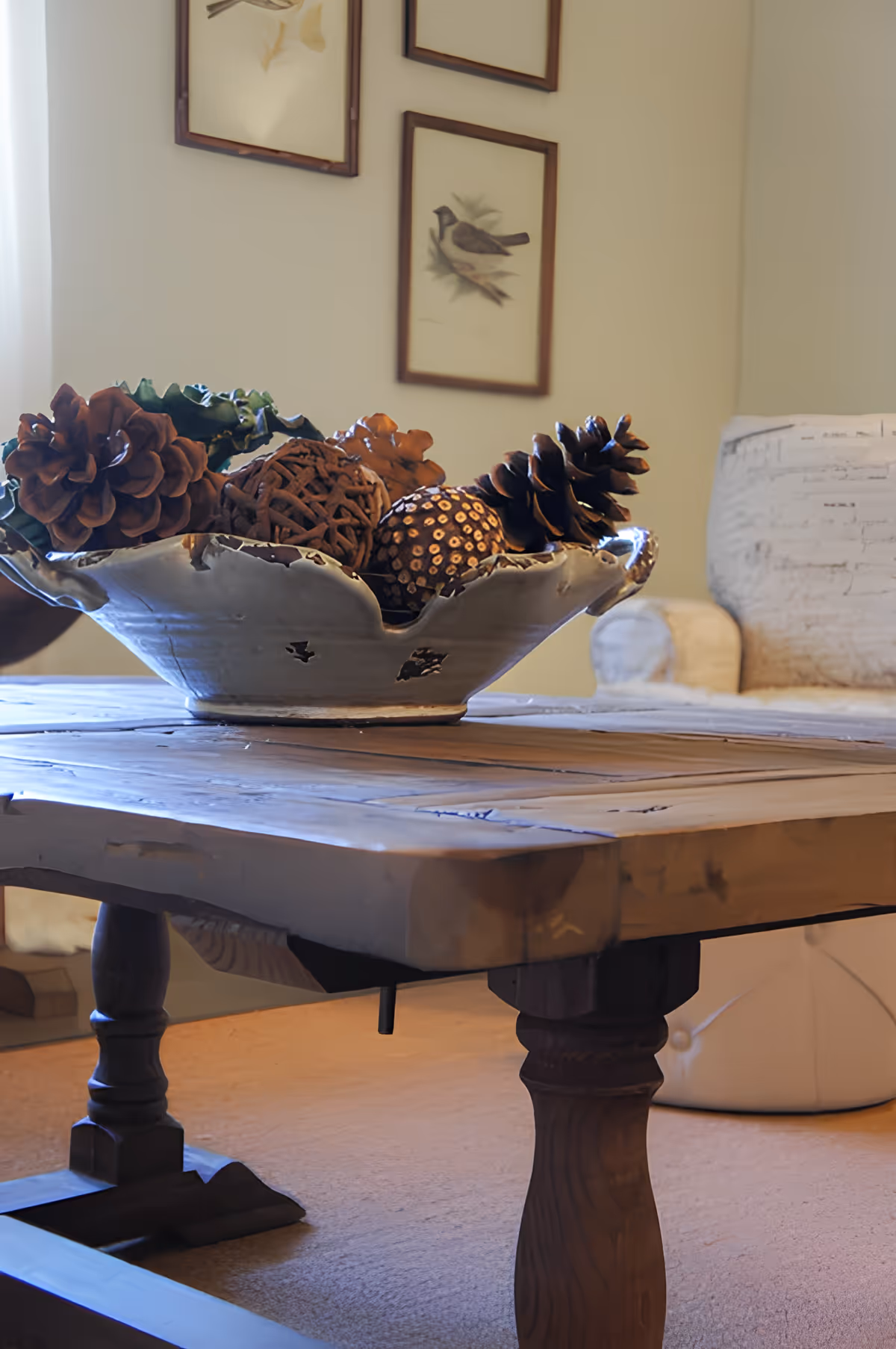 Close-up of a rustic wooden coffee table with a decorative ceramic bowl filled with pine cones and natural ornaments, set in a cozy living room with framed bird artwork on the wall and a cushioned armchair in the background.