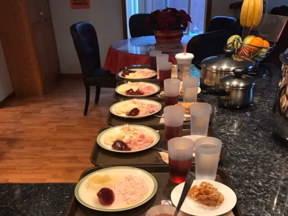 A kitchen counter with multiple trays lined up, each containing a plate of food with mashed potatoes, cranberry sauce, and a pink dish, along with cups of water and juice. In the background, there is a round dining table with a red tablecloth and a flower centerpiece, and some chairs around it. There are pots and a fruit basket on the counter as well.