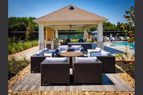 Outdoor seating area with dark wicker chairs and light cushions arranged around a circular fire pit table under a pavilion with a ceiling fan. In the background, there is a swimming pool with lounge chairs and greenery surrounding the area under a clear blue sky.