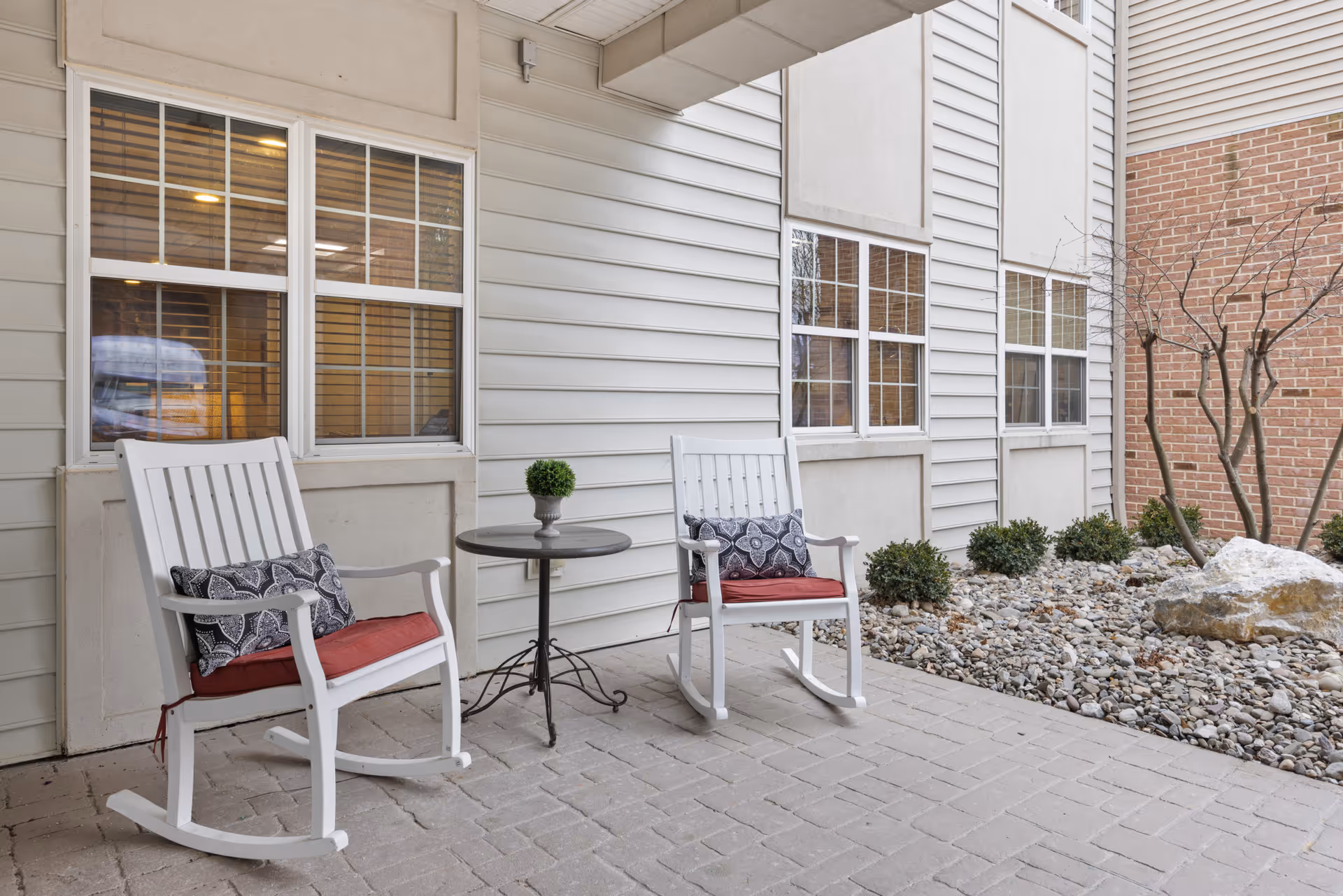 Outdoor patio area with two white rocking chairs featuring red cushions and patterned pillows, a small round table with a potted plant, and a landscaped garden with rocks and small bushes next to a building with white siding and brick walls.