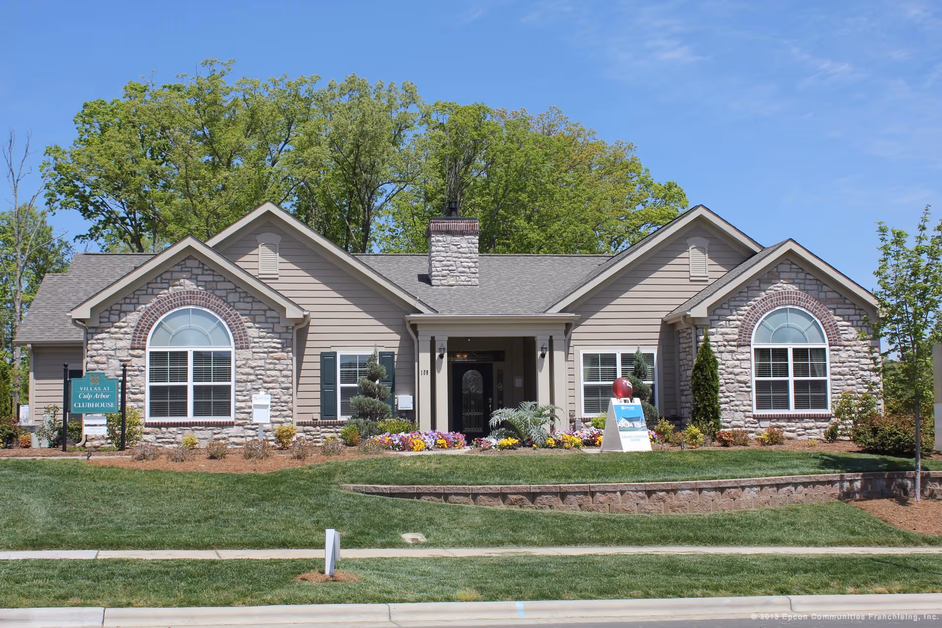 Front exterior view of a single-story clubhouse building with stone and siding facade, large arched windows, a central entrance with a covered porch, surrounded by landscaped greenery and flowers under a clear blue sky.