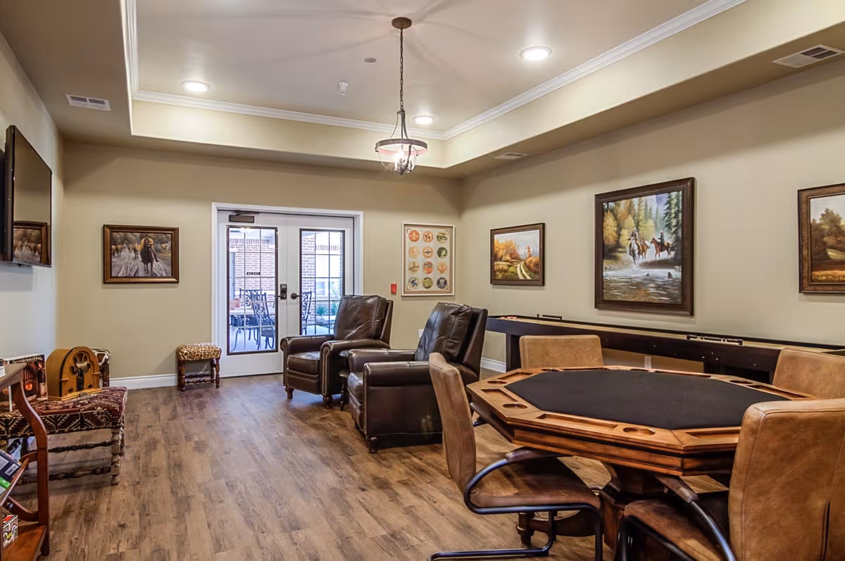 A cozy common room in an assisted living facility featuring a wooden octagonal game table with four chairs, two leather armchairs, a shuffleboard table along the wall, framed paintings, a flat-screen TV mounted on the wall, and double glass doors leading to an outdoor patio area.