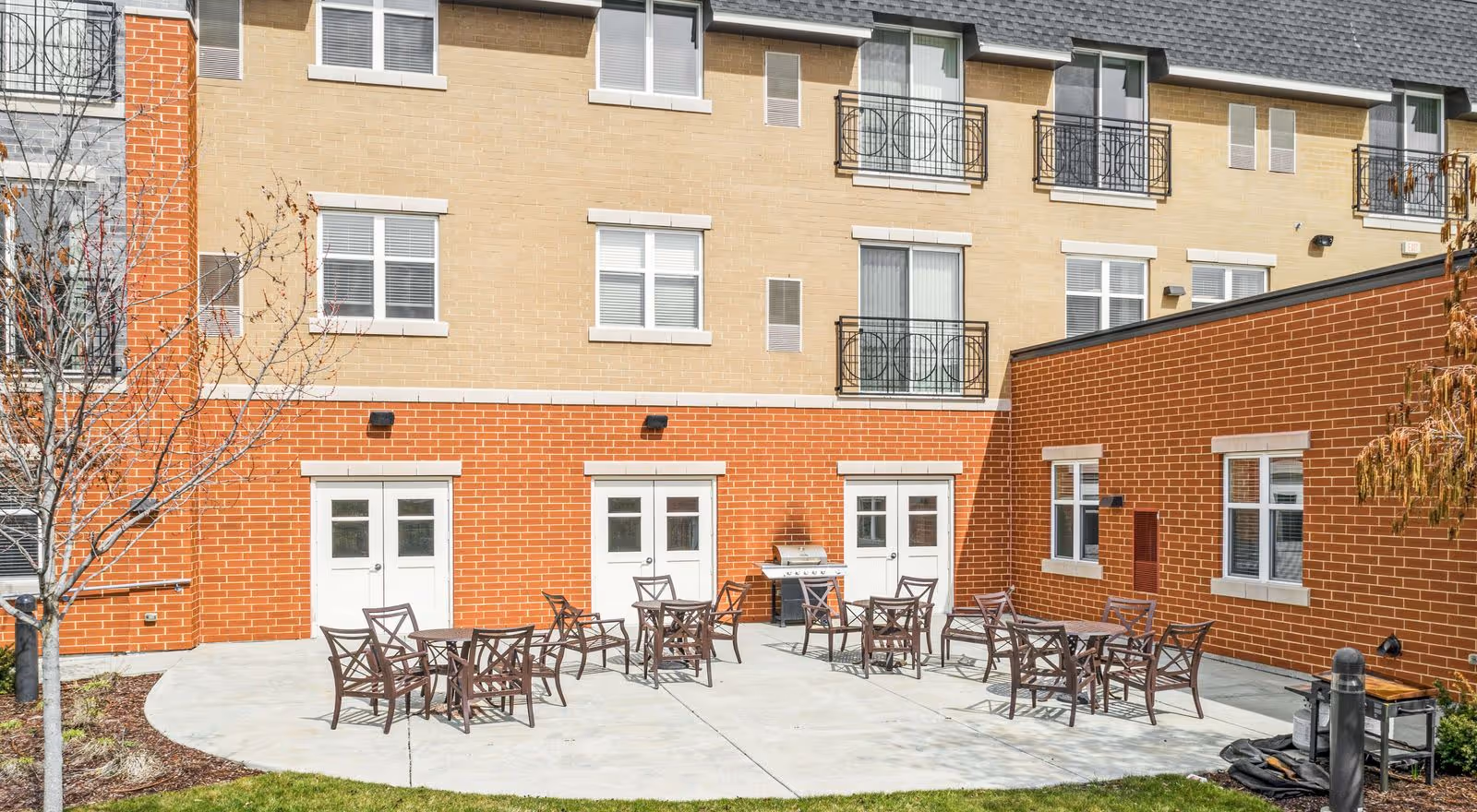 Outdoor patio area at a senior living facility with several metal tables and chairs arranged on a concrete surface. The building has a brick and beige exterior with multiple windows and small balconies. There is a grill and some landscaping with a tree and grass surrounding the patio.