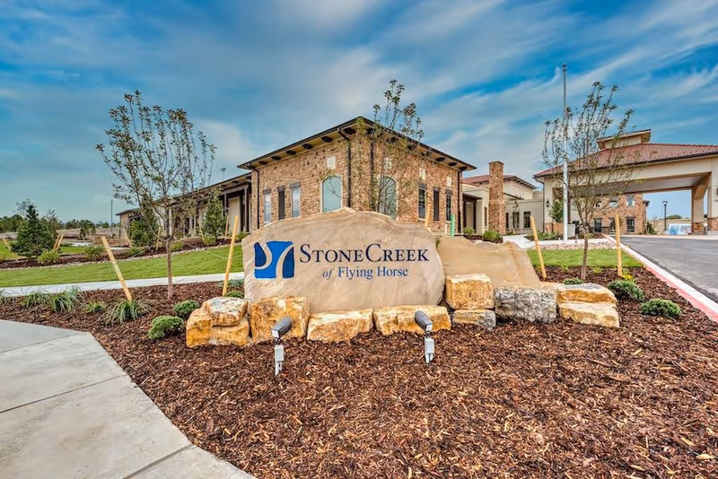 Exterior view of StoneCreek of Flying Horse Senior Living facility showing a large stone sign with the facility name in front of a landscaped area with small trees and shrubs, with the building and entrance visible in the background under a partly cloudy sky.