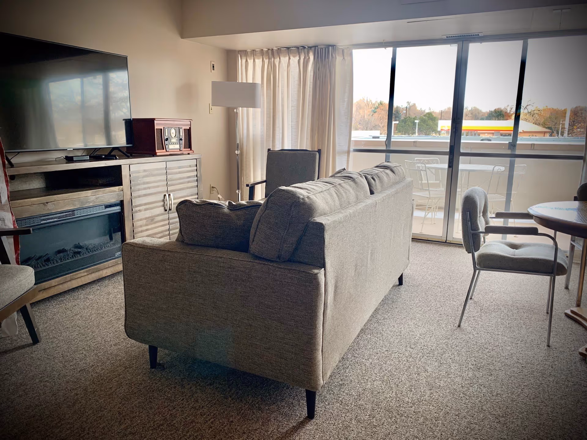 A living room with a gray sofa facing a large flat-screen TV on a wooden cabinet that also houses an electric fireplace. There is a floor lamp and an armchair near the window with closed curtains. Sliding glass doors lead to a balcony with outdoor furniture, and a round table with chairs is visible on the right side of the room.