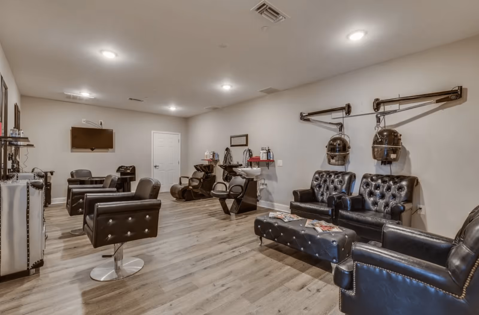 Interior view of a salon area in an assisted living facility featuring black leather chairs, hair washing stations, wall-mounted hair dryers, a TV, and a small table with salon products. The room has wood flooring and neutral-colored walls with ceiling lights.