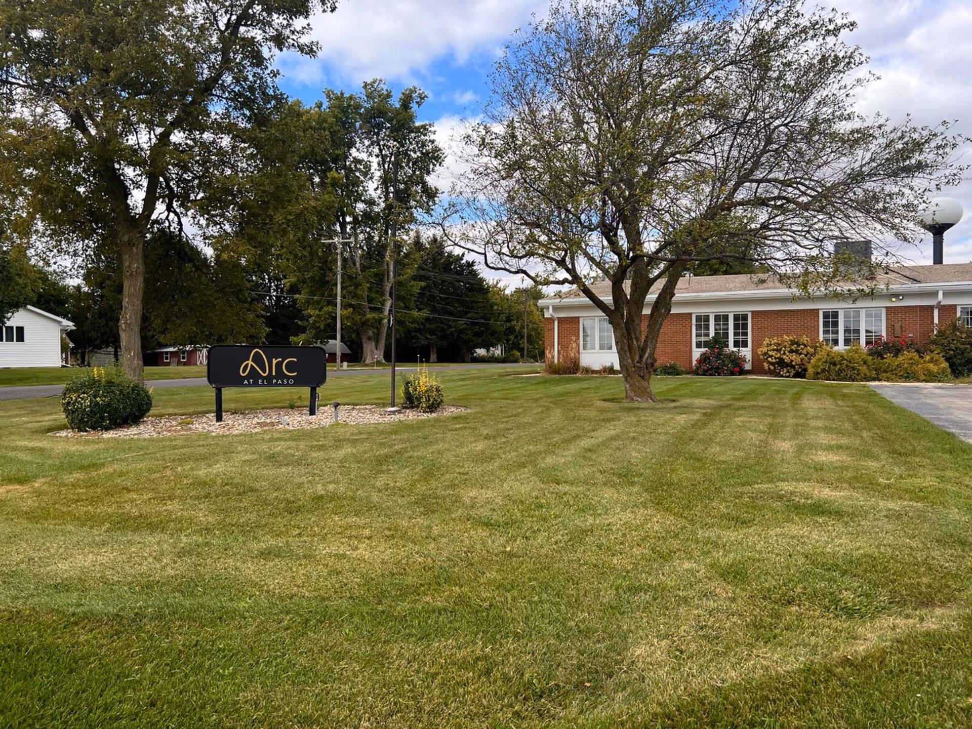 A grassy lawn with a few trees and a black sign that reads 'Arc at El Paso' in front of a single-story brick building with white-framed windows and some bushes. The sky is partly cloudy.