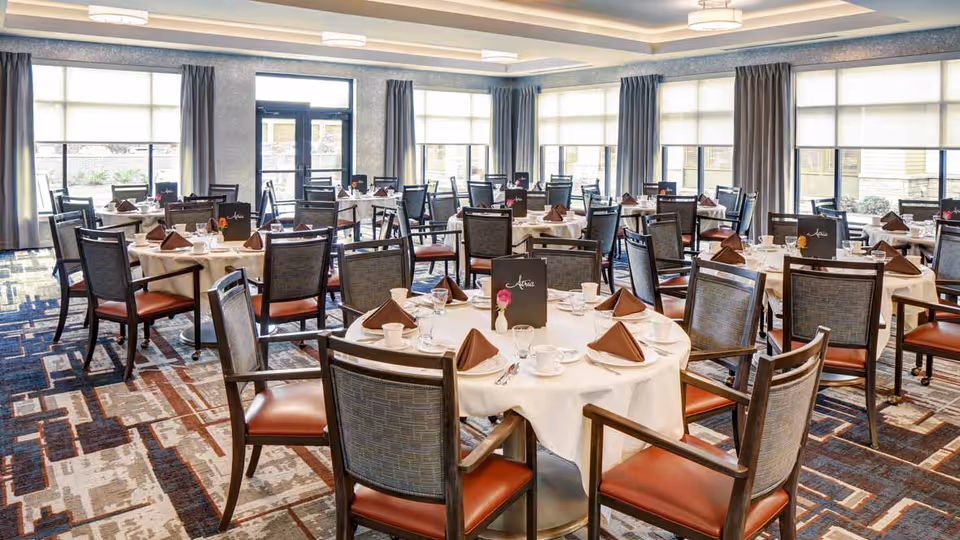 A spacious dining room with multiple round tables covered with white tablecloths, each set with brown folded napkins, white cups, glasses, and silverware. The room has large windows with gray curtains allowing natural light to fill the space. The carpet has a modern geometric pattern in shades of blue, brown, and beige.
