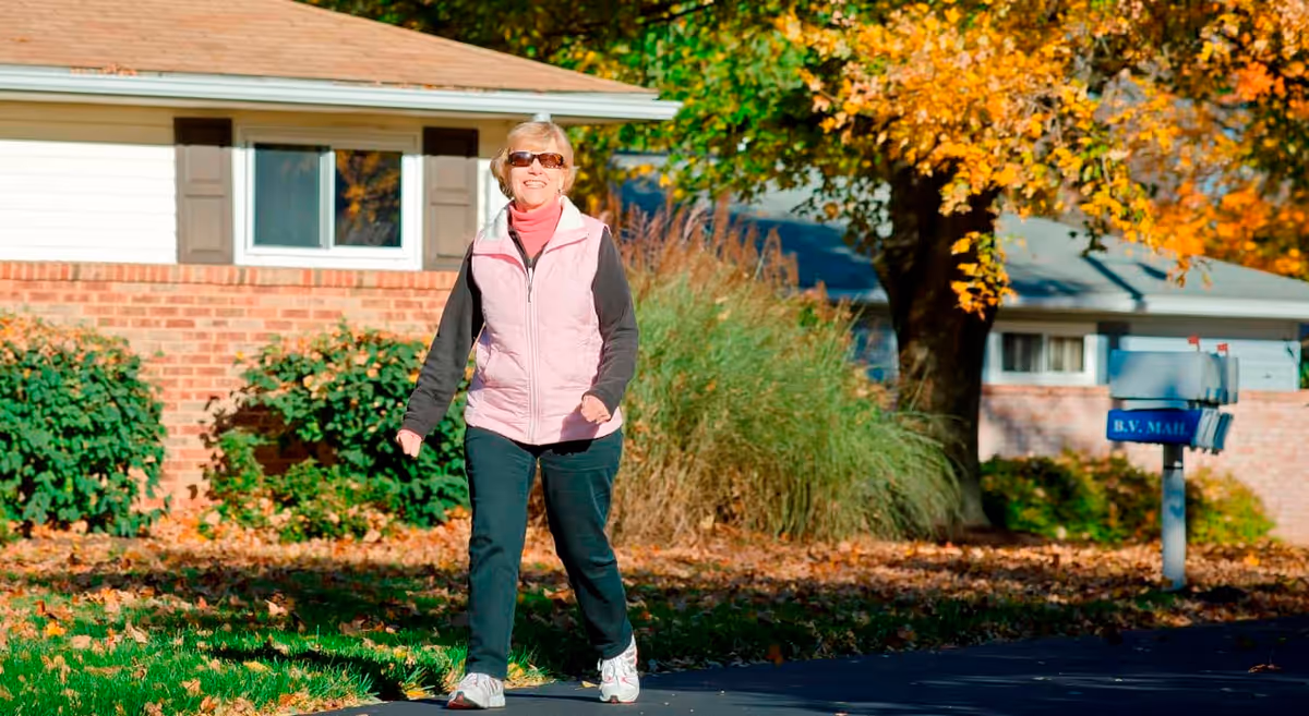 A woman wearing sunglasses and a pink vest walks on a sidewalk in front of a brick house and mailbox with autumn leaves on the ground.