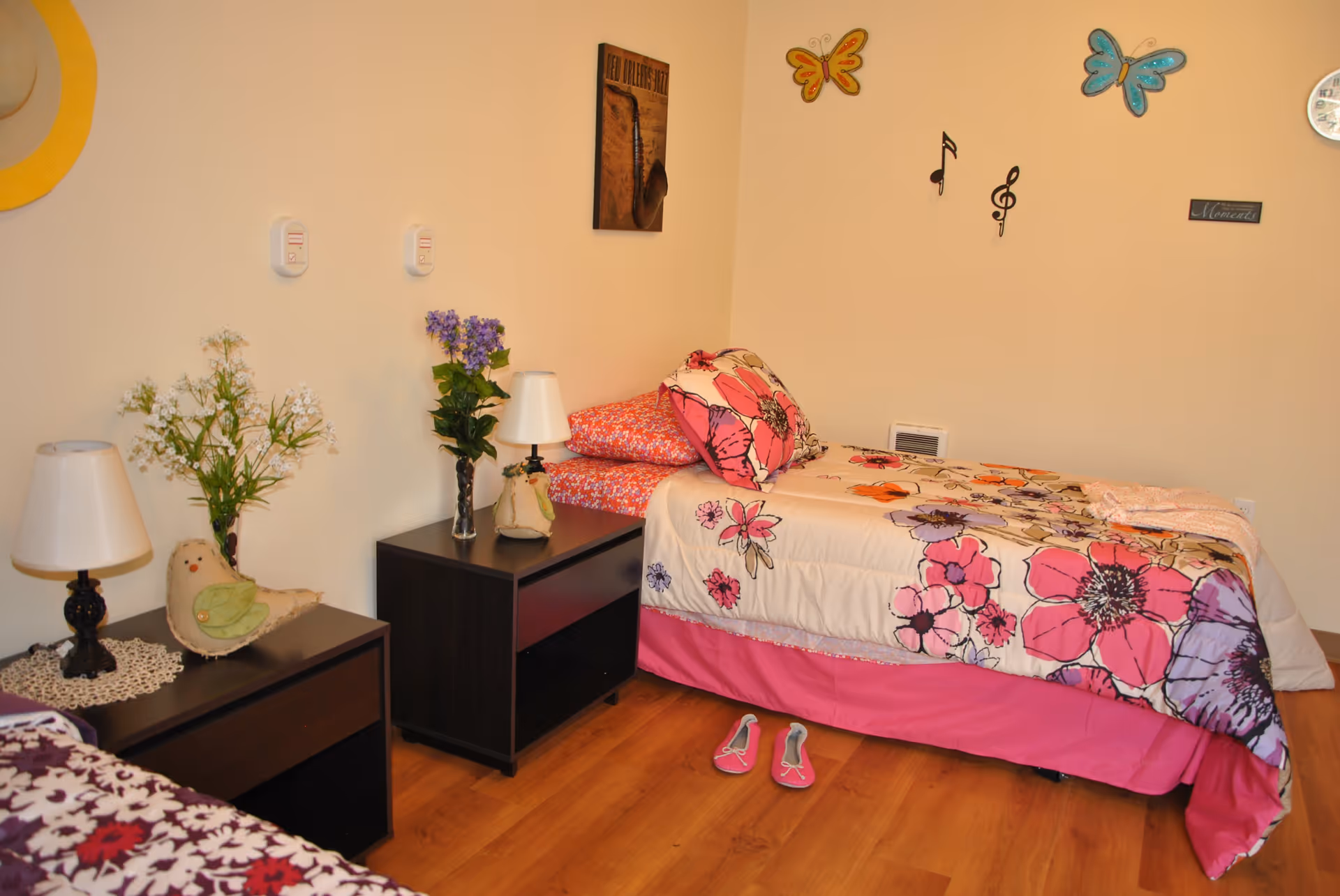 A cozy bedroom with a single bed covered in floral bedding in shades of pink, purple, and orange. The bed has matching pillows and a pink bed skirt. Next to the bed are two dark wooden nightstands, each with a lamp and decorative items including a vase with flowers and a small bird figurine. The walls are light-colored and decorated with butterfly and musical note wall hangings, a small framed picture, and a clock. The floor is wooden, and a pair of pink slippers is placed near the bed.