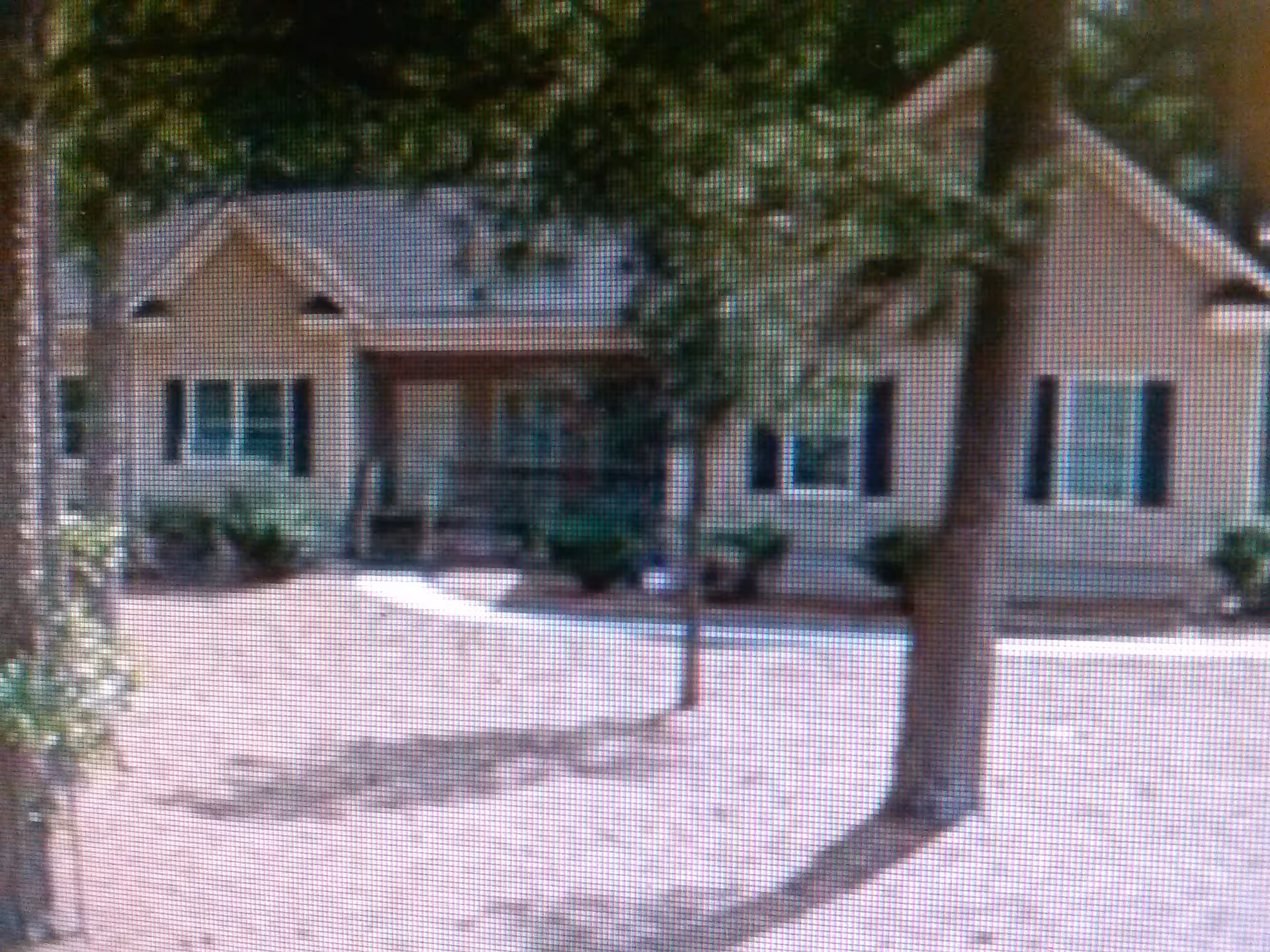Front view of a single-story house-like building with a porch, shuttered windows, trees and a bare yard in front.