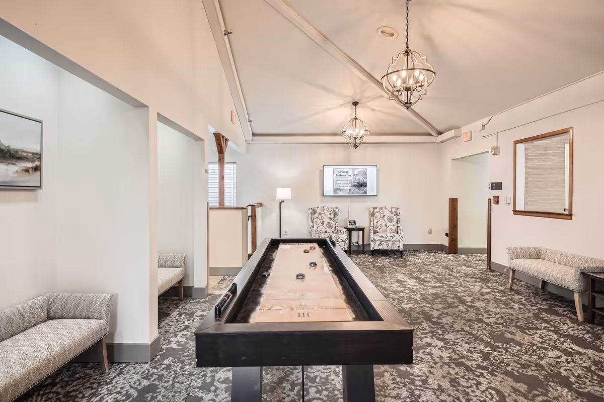 A bright and spacious common room with a shuffleboard table in the center, two patterned armchairs and a small table against the far wall, two cushioned benches along the side walls, a floor lamp, a wall-mounted TV, and two decorative hanging light fixtures on the ceiling.
