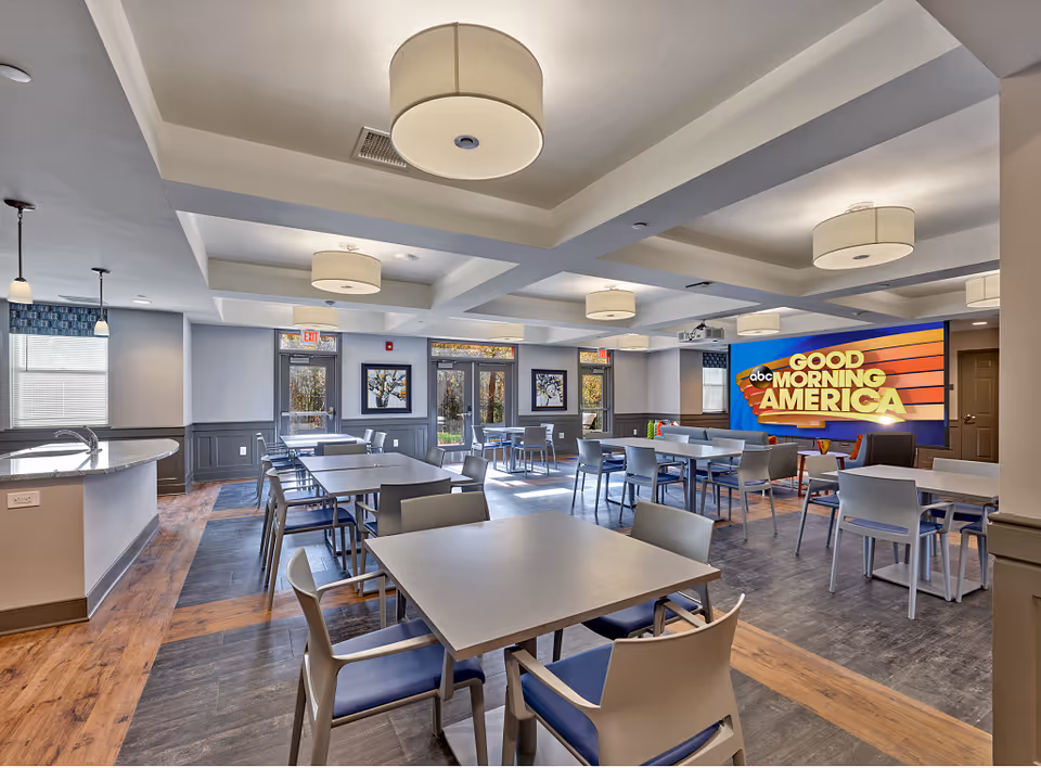 A bright and spacious dining area in a senior living facility with multiple tables and chairs arranged neatly. The room features modern ceiling lights, large windows letting in natural light, and a large screen on the wall displaying the Good Morning America logo. There is a kitchen counter with a sink on the left side and framed artwork on the walls.