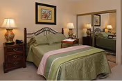 A bedroom with a bed covered in a green bedspread with pink and green stripes. There are two wooden nightstands on either side of the bed, each with a decorative lamp. A framed picture hangs on the wall above the bed. A large mirrored closet door is visible on the right side of the room.
