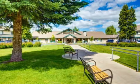 Outdoor view of a senior living facility with a paved walkway, two benches, green grass, trees, and the building entrance in the background under a partly cloudy sky.