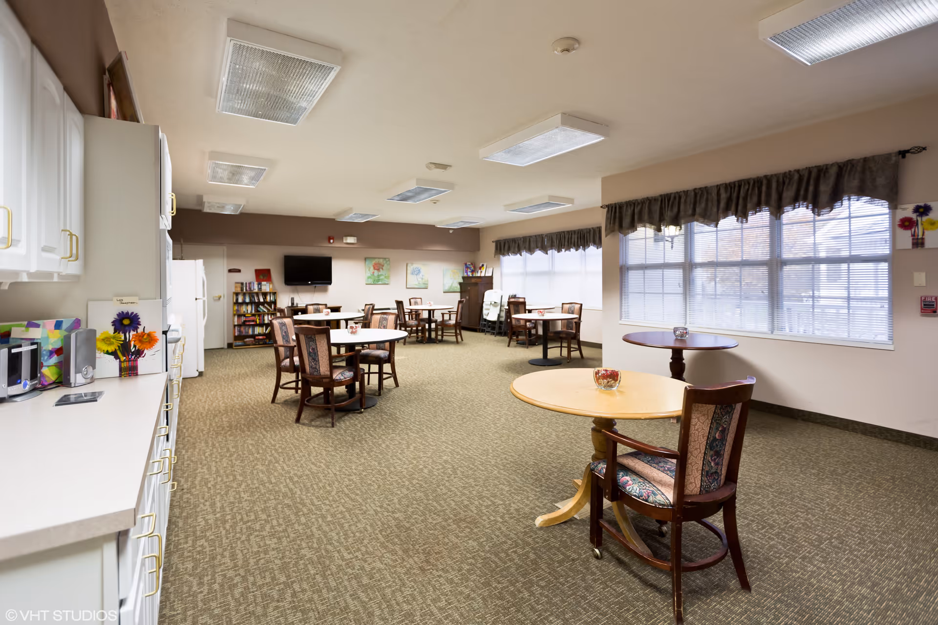 A spacious common area in a senior living facility with several round tables and chairs arranged throughout the room. There is a kitchenette area on the left side with white cabinets, a microwave, and a refrigerator. Large windows with valances allow natural light to fill the room. The walls are decorated with framed artwork, and there is a television mounted on the far wall above a bookshelf.