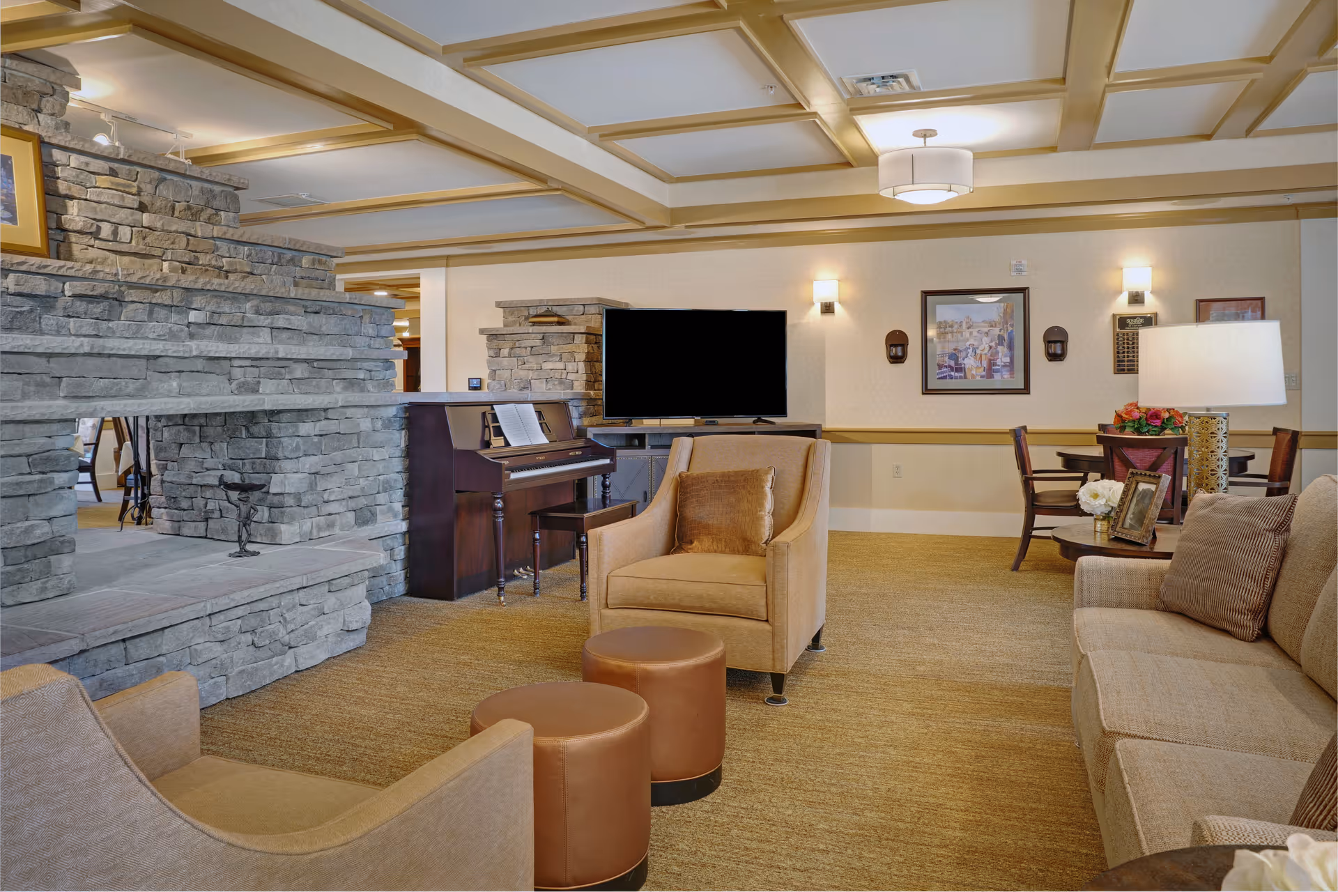 A cozy living room area in a senior living facility featuring a stone fireplace, a piano, a flat-screen TV, beige armchairs, a sofa, and a small round table with a lamp and flowers. The room has warm lighting and framed artwork on the walls.
