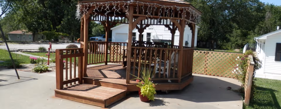 A wooden gazebo with steps, outdoor chairs, and a potted plant on a concrete patio beside light-colored buildings.