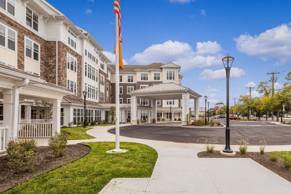 Exterior view of a senior living facility named Sunrise of New Dorp showing a multi-story building with a covered entrance, a flagpole with an American flag, landscaped green areas, sidewalks, street lamps, and a clear blue sky with some clouds.