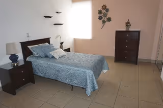 A simple bedroom with a single bed covered in a blue bedspread and pillows. There is a wooden nightstand with a lamp and a small plant next to the bed. On the opposite side of the room, there is a tall wooden dresser with a decorative item on top. The room has tiled flooring, a window with a blind, and a wall decoration with flowers.