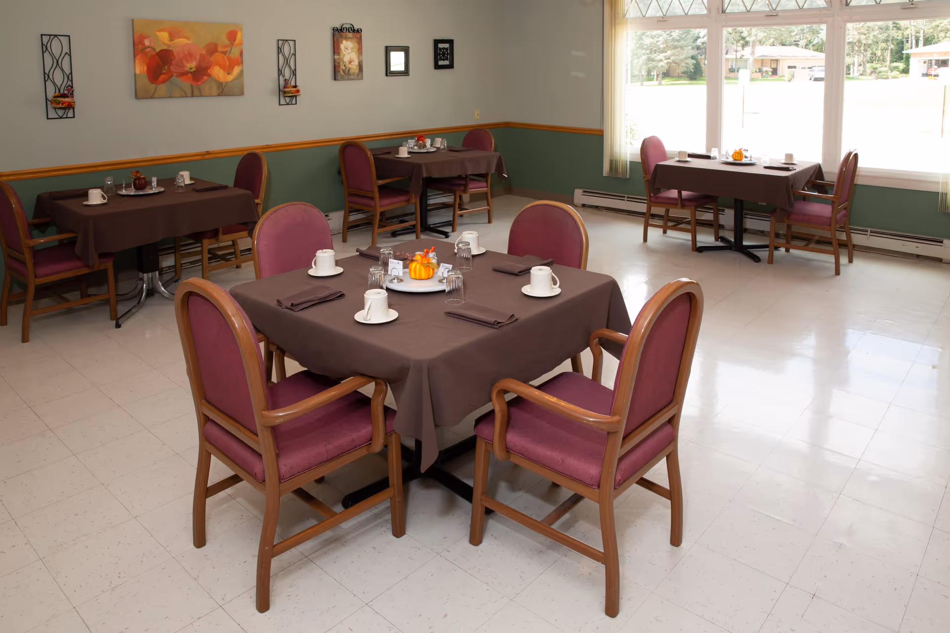 A dining room with several tables covered in brown tablecloths, each set with white cups, glasses, napkins, and small decorative pumpkins. The room has purple cushioned wooden chairs, light-colored tiled floor, green and beige walls with framed artwork, and large windows letting in natural light.