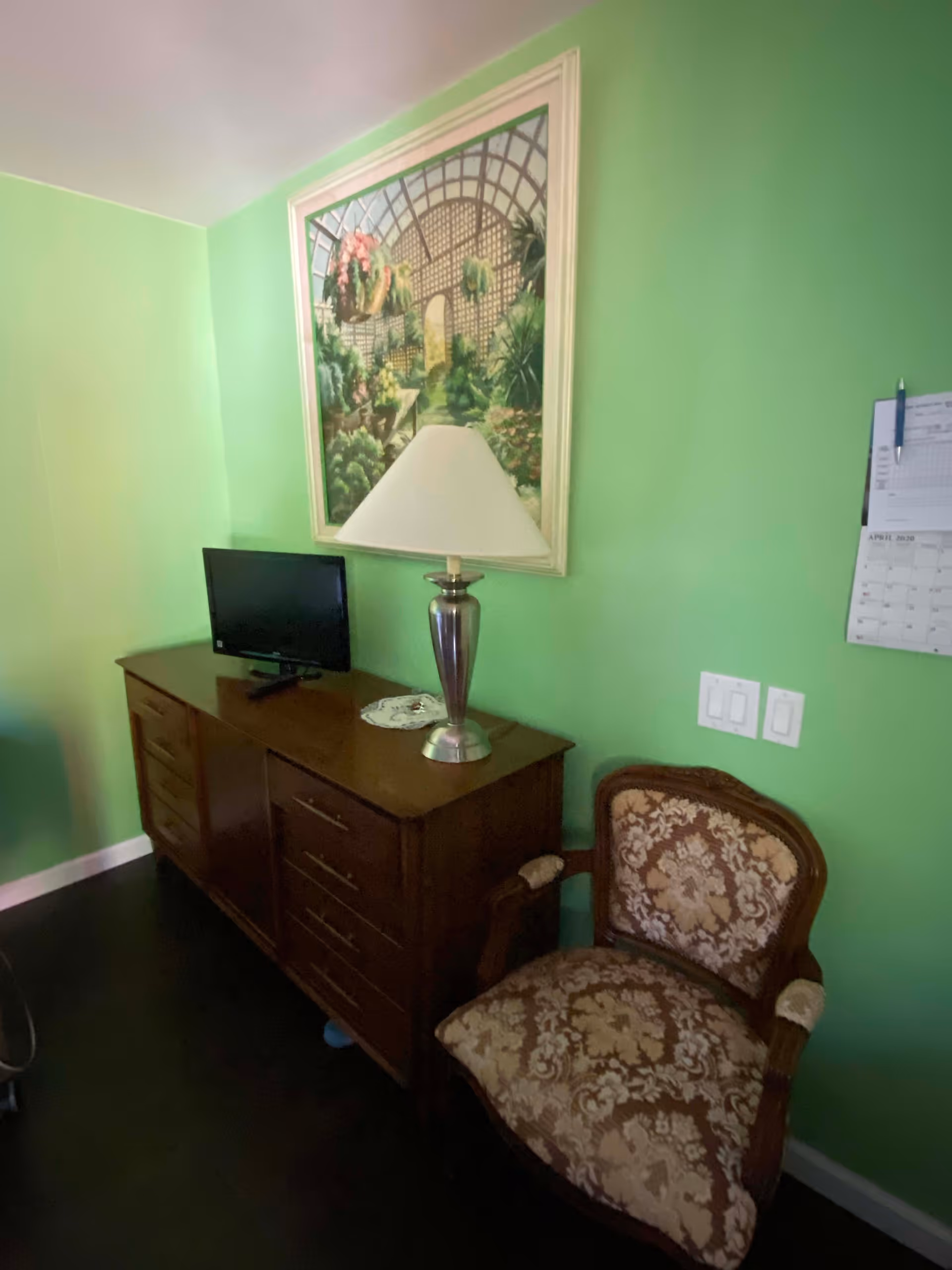 A room with light green walls featuring a wooden dresser with multiple drawers, a small flat-screen TV, and a silver table lamp with a white lampshade. Above the dresser hangs a framed painting of a garden scene. Next to the dresser is an upholstered armchair with a floral pattern. A calendar is partially visible on the wall to the right.